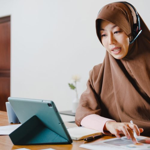 Asia muslim lady wear headphone using digital tablet talk to col Asia muslim lady wear headphone using digital tablet talk to colleagues about sale report in conference video call while working from home at kitchen. Social distancing, quarantine for corona virus.